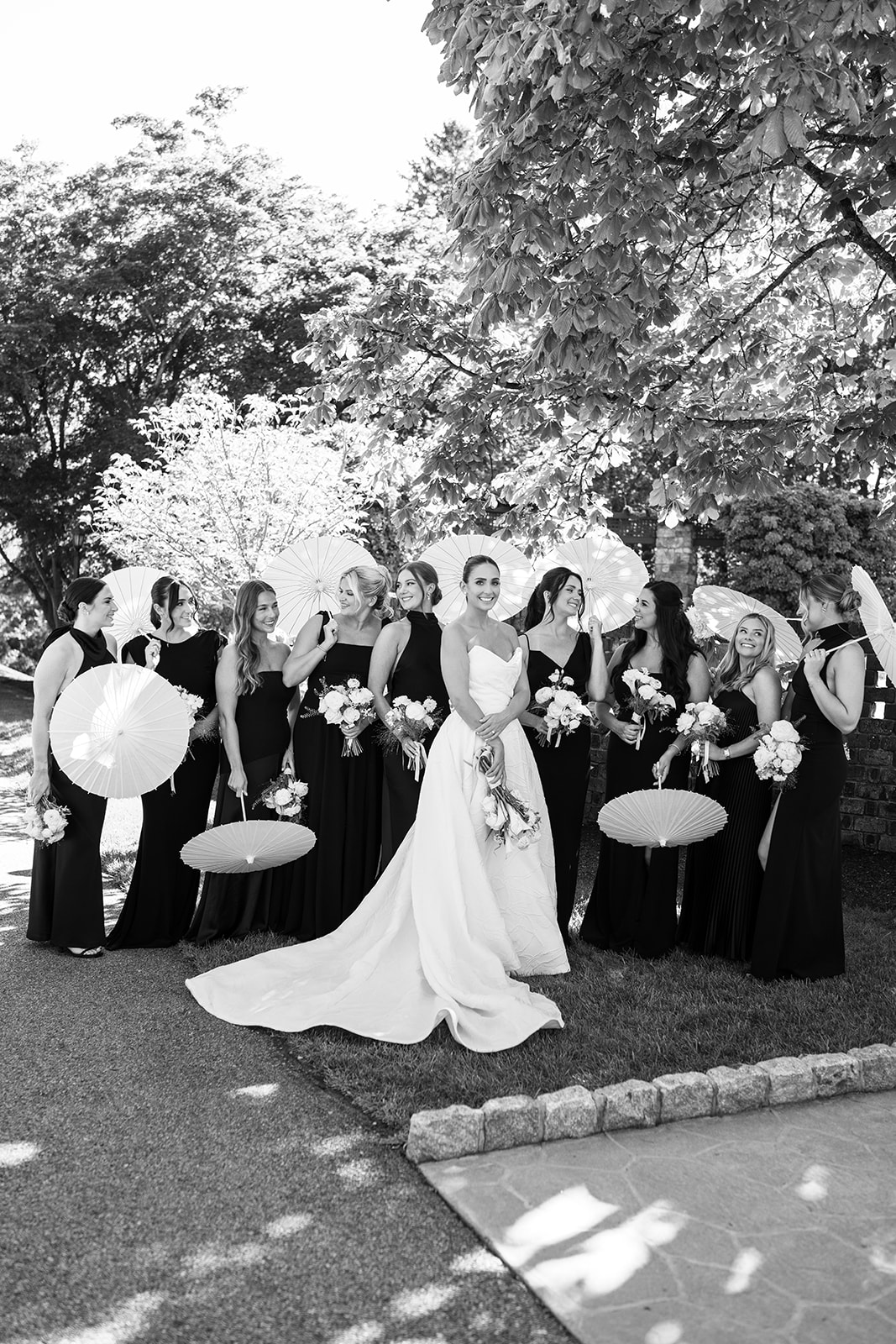Black and white film photo of the bride posing with her bridesmaids holding white paper parasols.